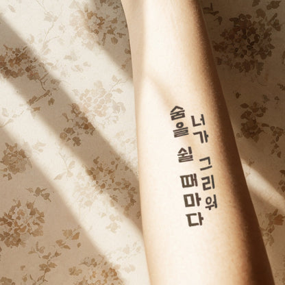 Forearm with a bold Korean script temporary tattoo, illuminated by soft sunlight filtering through window blinds onto a floral-patterned surface.