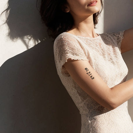 A woman in a lace dress, standing in a sunlit space, with a Korean phrase temporary tattoo prominently displayed on her upper arm.