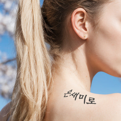 Profile view of a woman with a ponytail showcasing a temporary Korean tattoo in Hangul script on her neck, with a backdrop of blue sky and cherry blossoms.