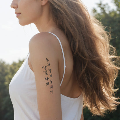 Side profile of a woman in a white dress with flowing hair, showcasing a column of traditional Korean temporary tattoos in Hangul script on her upper arm, with a natural landscape in the background.