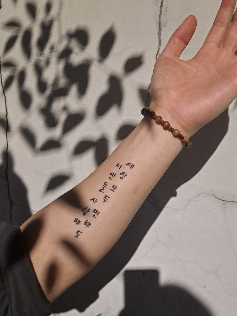 An arm extended against a textured wall with leafy shadows, featuring a line of Hangul script as a temporary tattoo, complemented by a wooden bead bracelet.