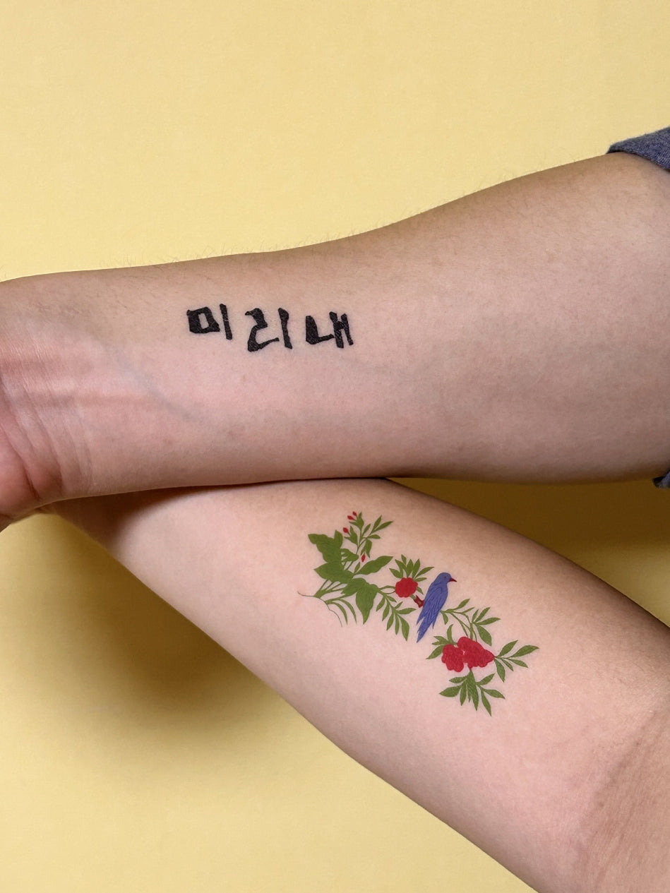 An arm with a Hangul script temporary tattoo placed on the inner forearm, above a colorful botanical design tattoo, against a yellow background.