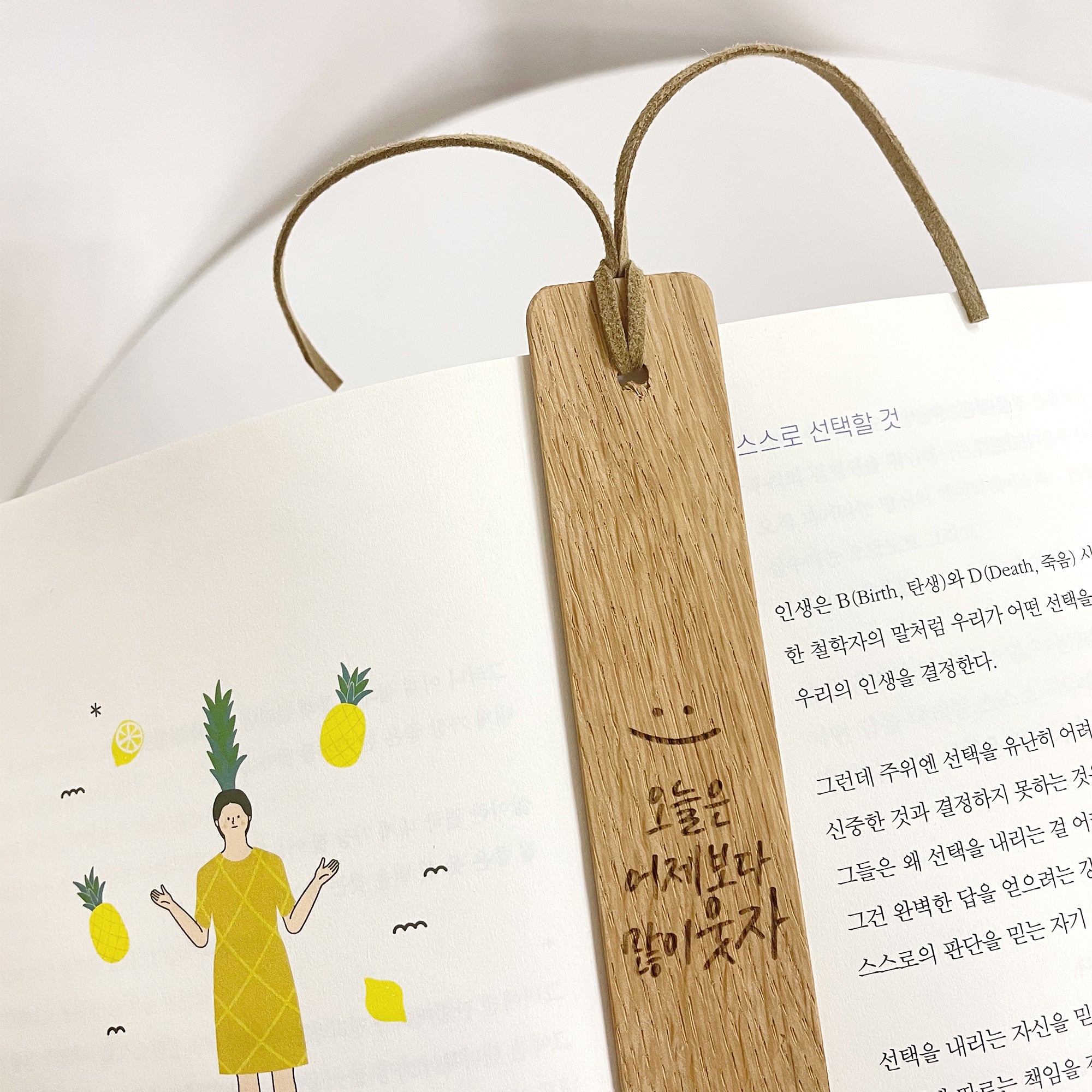 Handmade wooden bookmark with a simple smiling face illustration and Korean calligraphy, marking a page in a book adorned with pineapple graphics—a unique personalized gift.