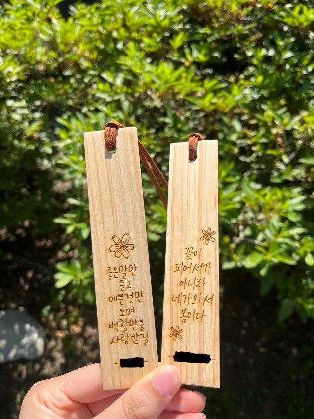Two wooden bookmarks with personalized Korean calligraphy and small flower engravings, held up against a green shrubbery background.