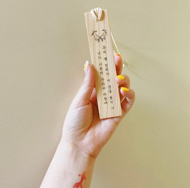 A hand holding a handmade wooden bookmark with Korean calligraphy and a heart and arrow illustration, complemented by the person's bright yellow nail polish and a whimsical temporary tattoo on the wrist.