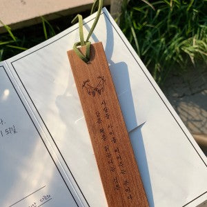 Handmade wooden bookmark with engraved Korean calligraphy and a drawing of a cat's face, resting on an open book's page in sunlight.